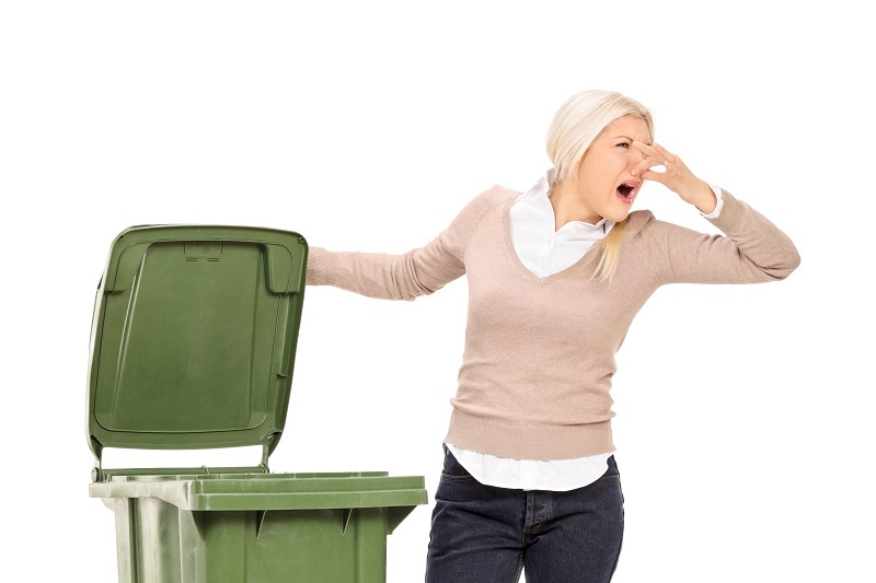 A woman opens a trashcan and is holding her nose because of the smell